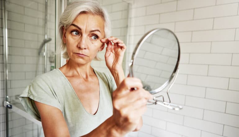 A woman touches her cheek as she looks in a hand mirror at her face.