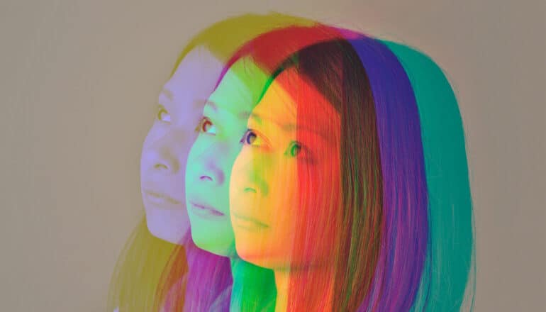 A young woman looks into the distance as many faint but colorful images of her face blend together.