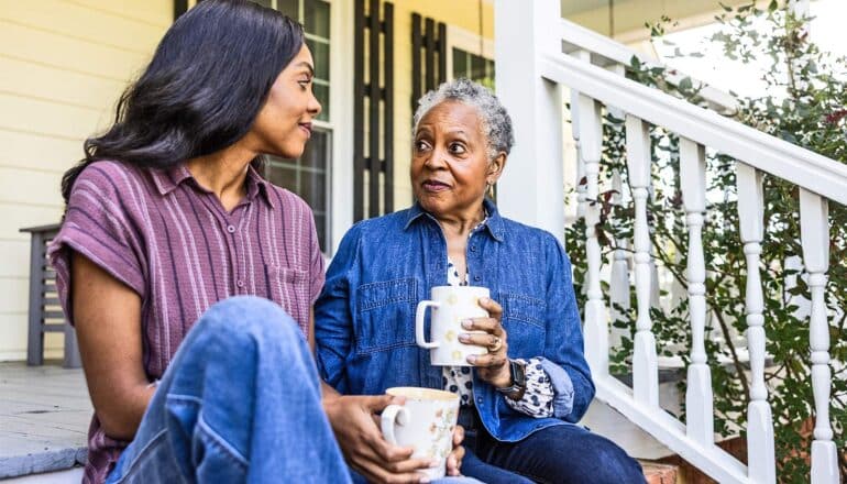 A mother and daughter sit sipping coffee on a porch.