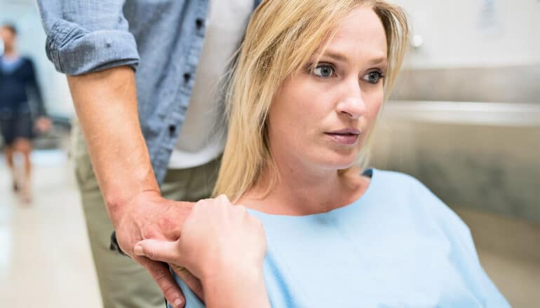 A woman in a wheelchair with a nervous look on her face is pushed down a hospital hallway by a man whose hand she's holding.
