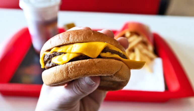 A first-person perspective of a person holding a cheeseburger with a tray holding fries and a soda in the background.