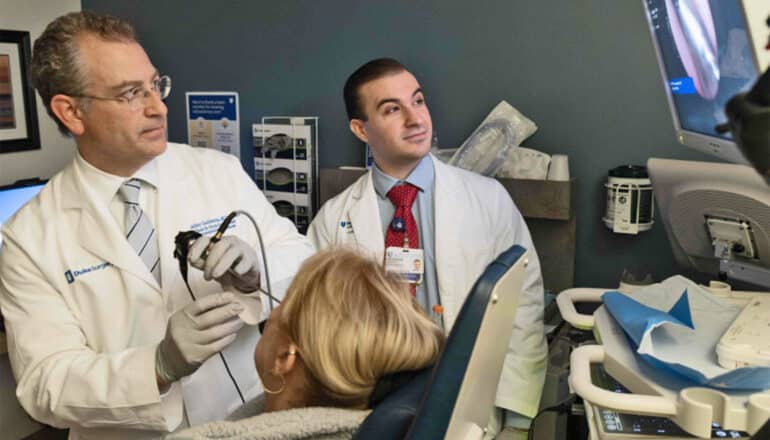 A man in a white coat uses a nasal swab test on a woman in a chair while he and another man in a white coat look up at a screen showing her nasal cavity.