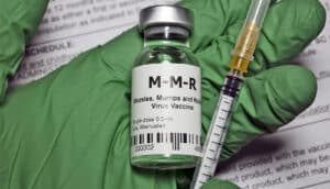A doctor wearing a green glove holds a vial of the MMR vaccine and a syringe.
