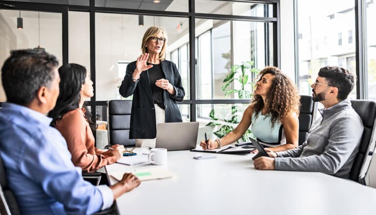 A manager speaks to her employees sitting around a conference table.