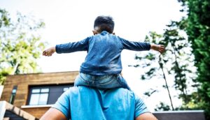 A young child with arms outstretched sits on a father's shoulders.