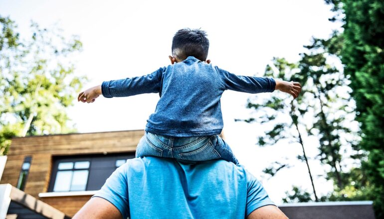 A young child with arms outstretched sits on a father's shoulders.