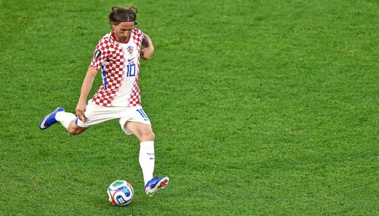 Modric lifts his right leg to kick the soccer ball on a deep green field.