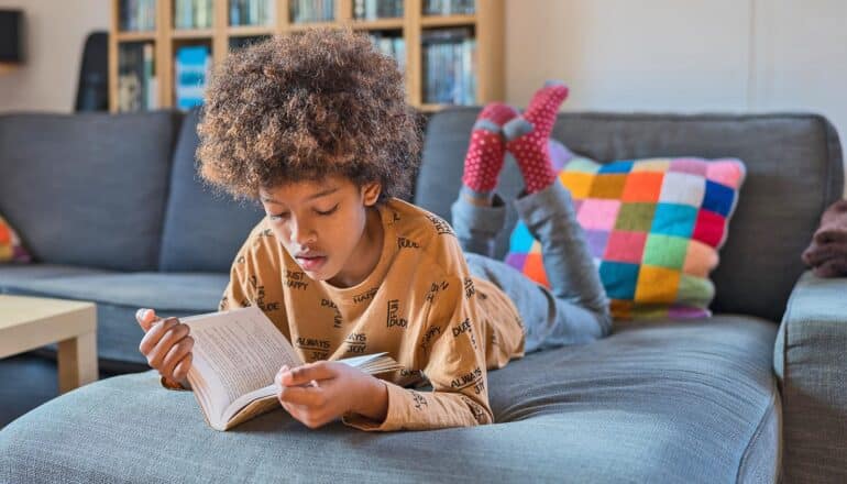 A young boy reads alone while laying on a couch.