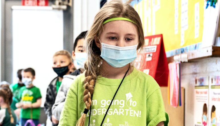 A young girl wearing a medical mask stands at the front of a line of young school kids in a classroom.